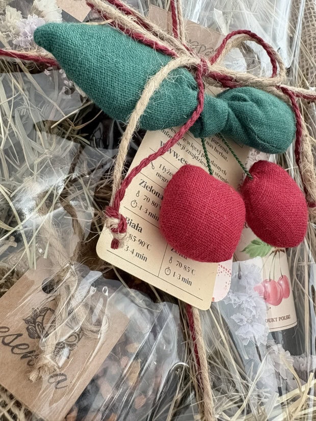 Zbliżenie na ręcznie szytą zawieszkę w kształcie wiśni przy zestawie prezentowym Wiśniowa Słodycz. W tle naturalne dekoracje z juty, sznurka i suszonych kwiatów.