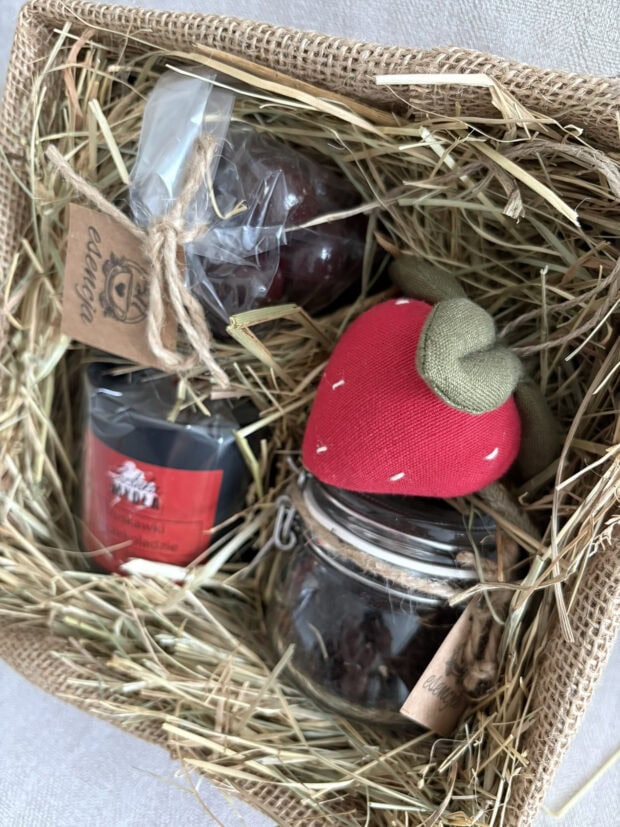 Zestaw prezentowy Truskawkowa Rozkosz zapakowany w jutę i celofan, ozdobiony ręcznie szytą zawieszką w kształcie truskawki. W środku świeca sojowa, aromatyczna herbata i truskawki w czekoladzie.