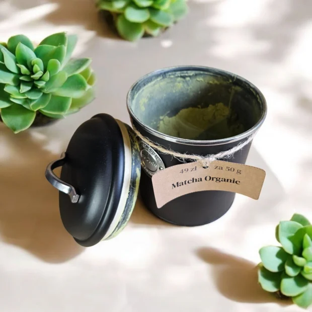 Czarna puszka z organiczną matchą, otwarta, ustawiona na tle dekoracyjnych sukulentów.