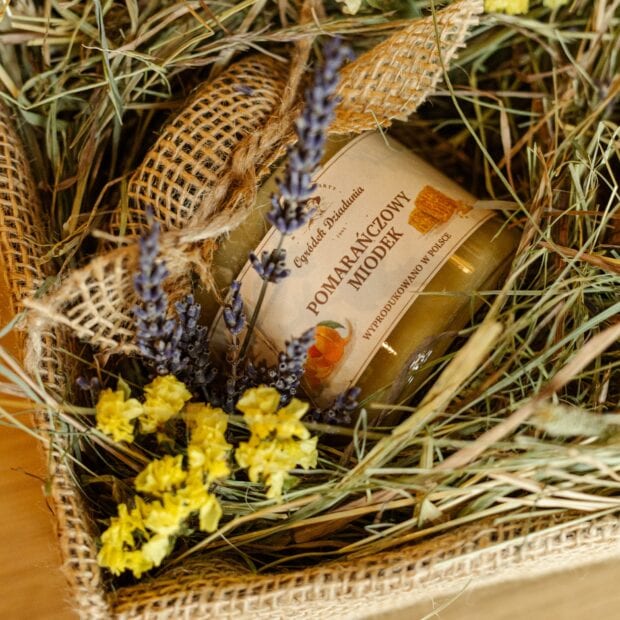 Miodek pomarańczowy z zestawu prezentowego, otoczony naturalnymi dekoracjami - siankiem i kwiatami.