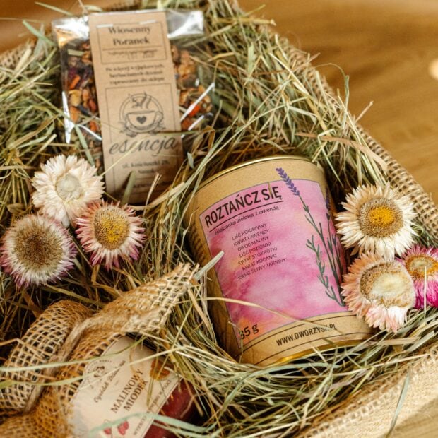 Herbaty i puszka ziół Dworzysk w zestawie prezentowym, otoczone naturalnymi dodatkami - siankiem i kwiatami.