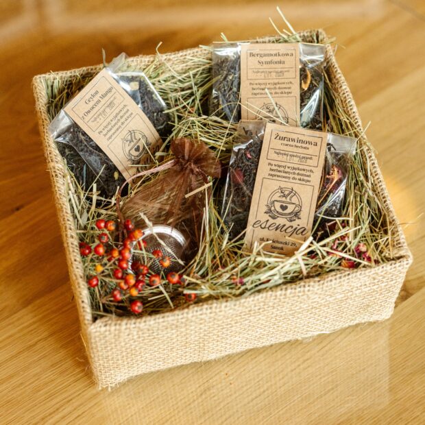 Herbaty w transparentnych torebkach umieszczone w pudełku ozdobnym, otoczone siankiem i kwiatami, wraz z zaparzaczem.