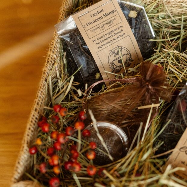 Kadr z pudełkiem prezentowym z herbatami i zaparzaczem, ozdobione siankiem i kwiatami.