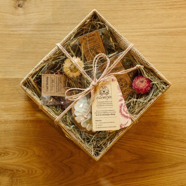 Pudełko prezentowe, zawiązane sznurkiem i zawinięte celofanem z torebkami transparentnymi herbaty w środku i pierniczkiem, ozdobione siankiem i kwiatami w środku.