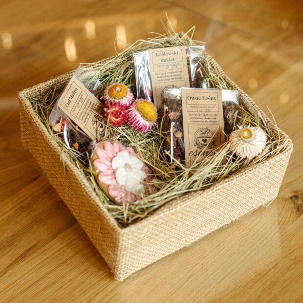 Pudełko jutowe wypełnione siankiem i kwiatami z suszami owocowymi w transparentnych torebkach i pierniczkiem.