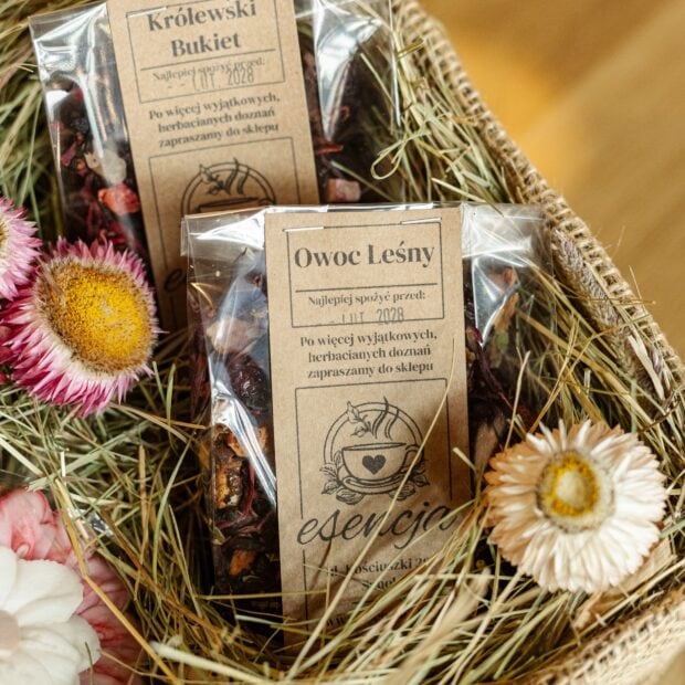 Dwie paczuszki suszu owocowego w pudełku, otoczone siankiem i kwiatami.