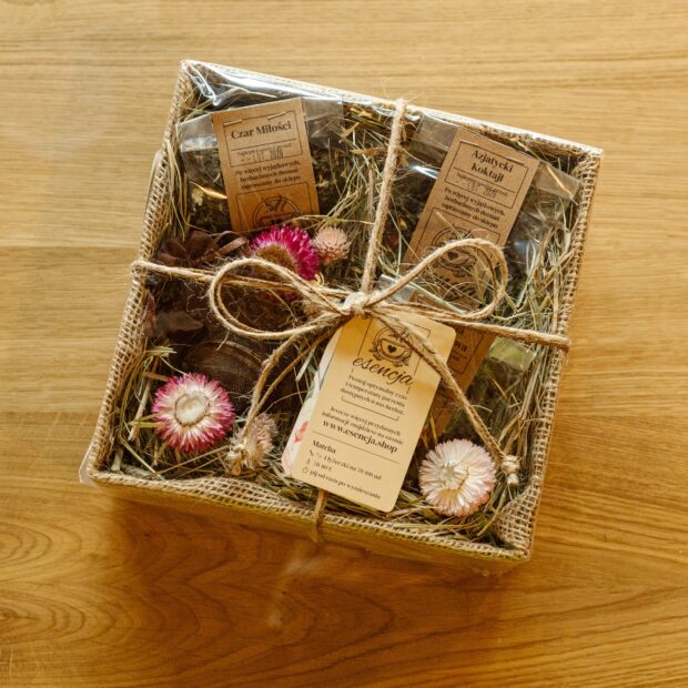 Pudełko prezentowe, zawiązane sznurkiem, opatrzone etykietą, w środku widoczne kwiaty i sianko oraz herbaty spakowane w transparentne torebki.