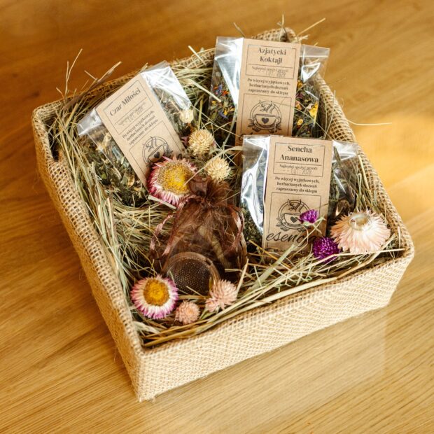 Ozdobne pudełko z herbatami w transparentnych torebkach w otoczeniu sianka i kwiatów, z zaparzaczem w woreczku.