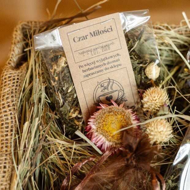 Zielona herbata w transparentnej torebce otoczona siankiem i kwiatami.