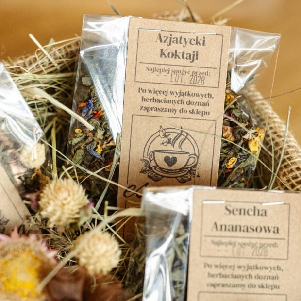 Dwie torebki transparentne z herbatą w pudełku jutowym, otoczone siankiem i kwiatami.